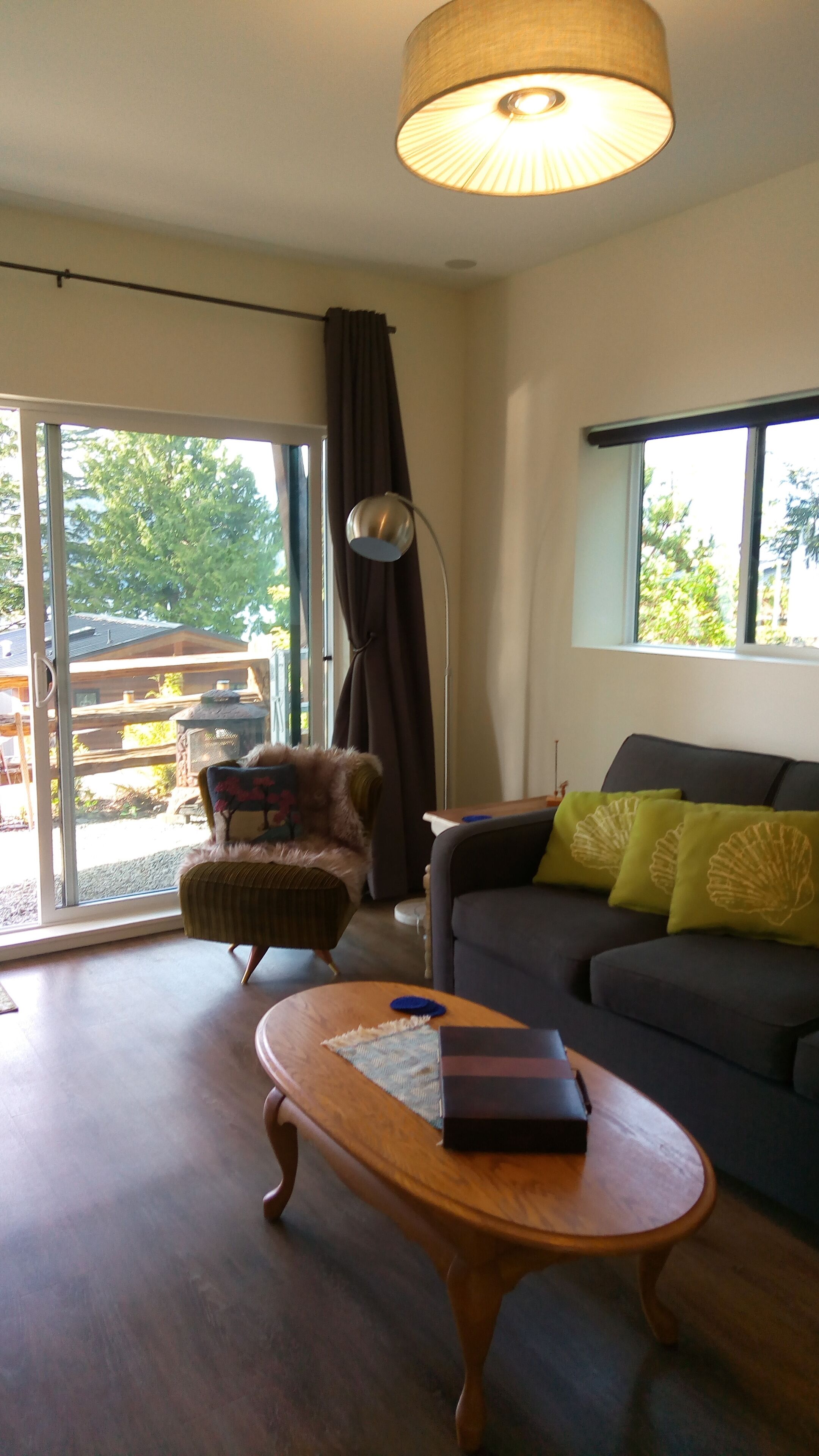 A modern living room with a sofa, armchair, and a coffee table. There is a large window with curtains, a floor lamp, and a view of the outdoors.