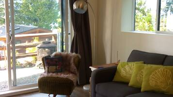 A modern living room with a sofa, armchair, and a coffee table. There is a large window with curtains, a floor lamp, and a view of the outdoors.