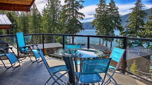 Un balcon avec une table et des chaises en verre, donnant sur un lac et des montagnes boisées.