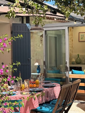 An outdoor dining area with a table set for two, a blue chair, and a glass door leading to a room with a bed and a lamp. - HOUSE OF 3 BEDROOMS 10 MIN WALK FROM THE CENTER OF SAINT REMY DE PROVENCE (Saint-Rémy-de-Provence)