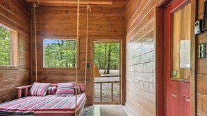 Interior de una cabaña de madera con una cama, una puerta roja y una ventana con vistas a un bosque.