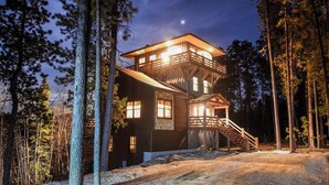 A multi-story wooden cabin with a balcony, a staircase, and a gravel driveway surrounded by trees.