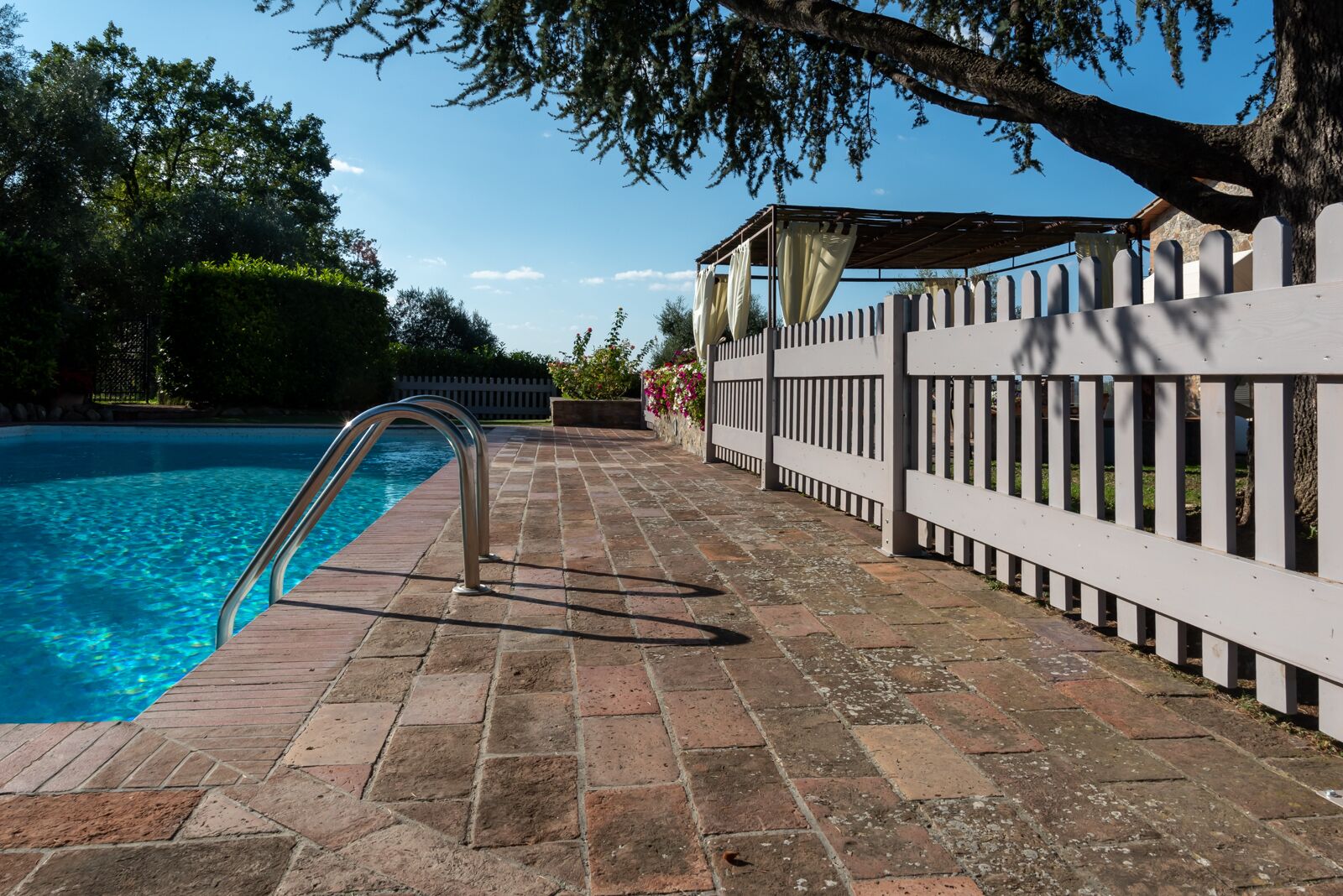 A swimming pool with a metal ladder, surrounded by a brick walkway and a white picket fence.