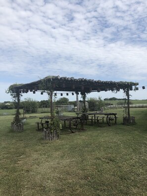 A rustic outdoor seating area with a wooden pergola, picnic tables, and hanging lights. - “3-Shy Ranch 40-Acre Texas Retreat Sleeps Up to 24 Near Tarleton” (Stephenville)