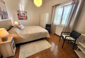 Ein Schlafzimmer mit einem Bett, einem Sessel, einem Kleiderschrank, einem Schreibtisch mit Lampe und einem Bild an der Wand.