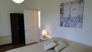 Ein Schlafzimmer mit einem Bett, einem Nachttisch, einer Lampe, einer Tür und einem Poster von Paris an der Wand.
