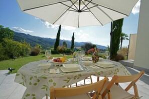 Eettafel buiten gedekt met borden, glazen en een fruitschaal, onder een grote parasol, met een prachtig uitzicht op de bergen op de achtergrond.