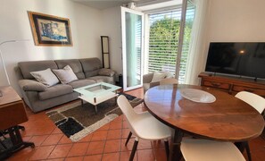 Ein Wohnzimmer mit einer Couch, einem Couchtisch, einem Esstisch, Stühlen, einem Fernseher und einer Schiebetür zu einem Balkon.
