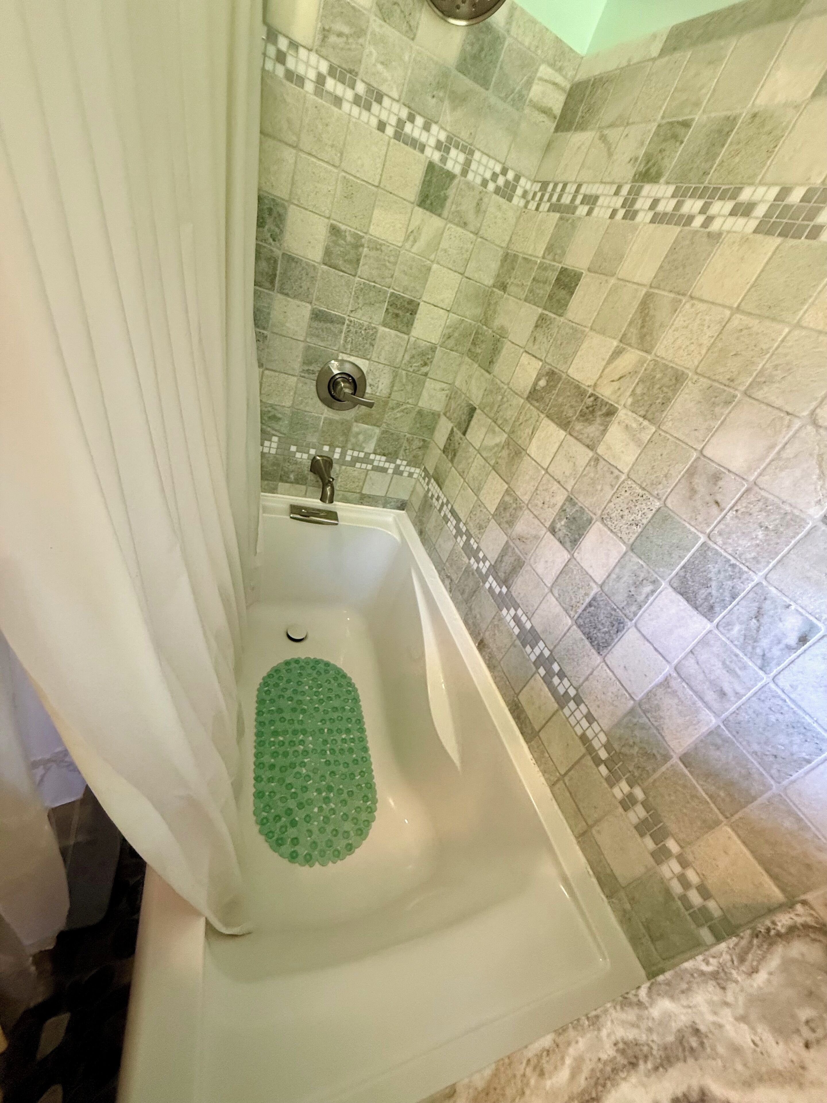Una bañera con un tapete de baño verde, un cabezal de ducha y una pared de azulejos con un diseño a cuadros.
