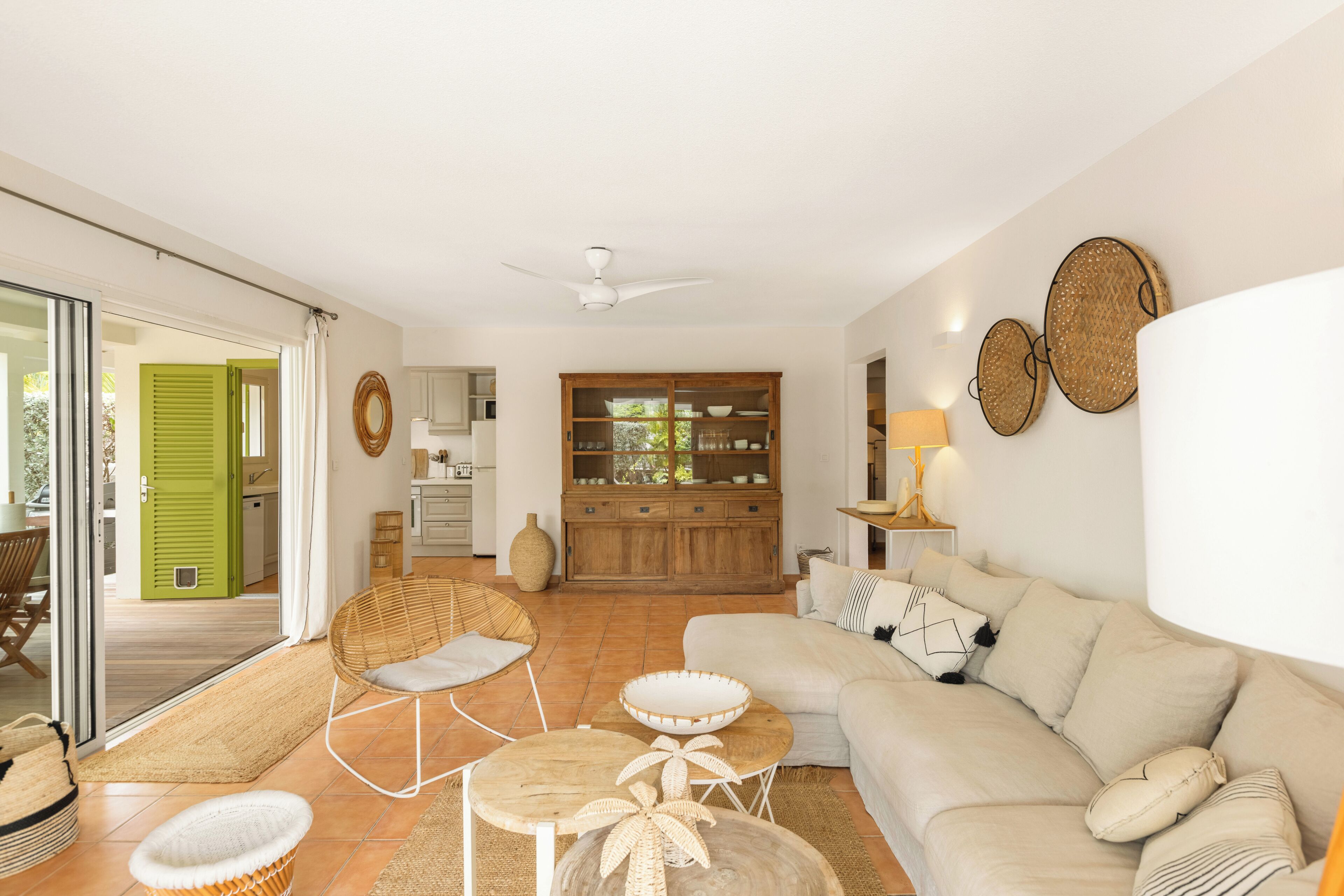 A living room with a sofa, a dining table, a wicker chair, and a kitchen in the background.