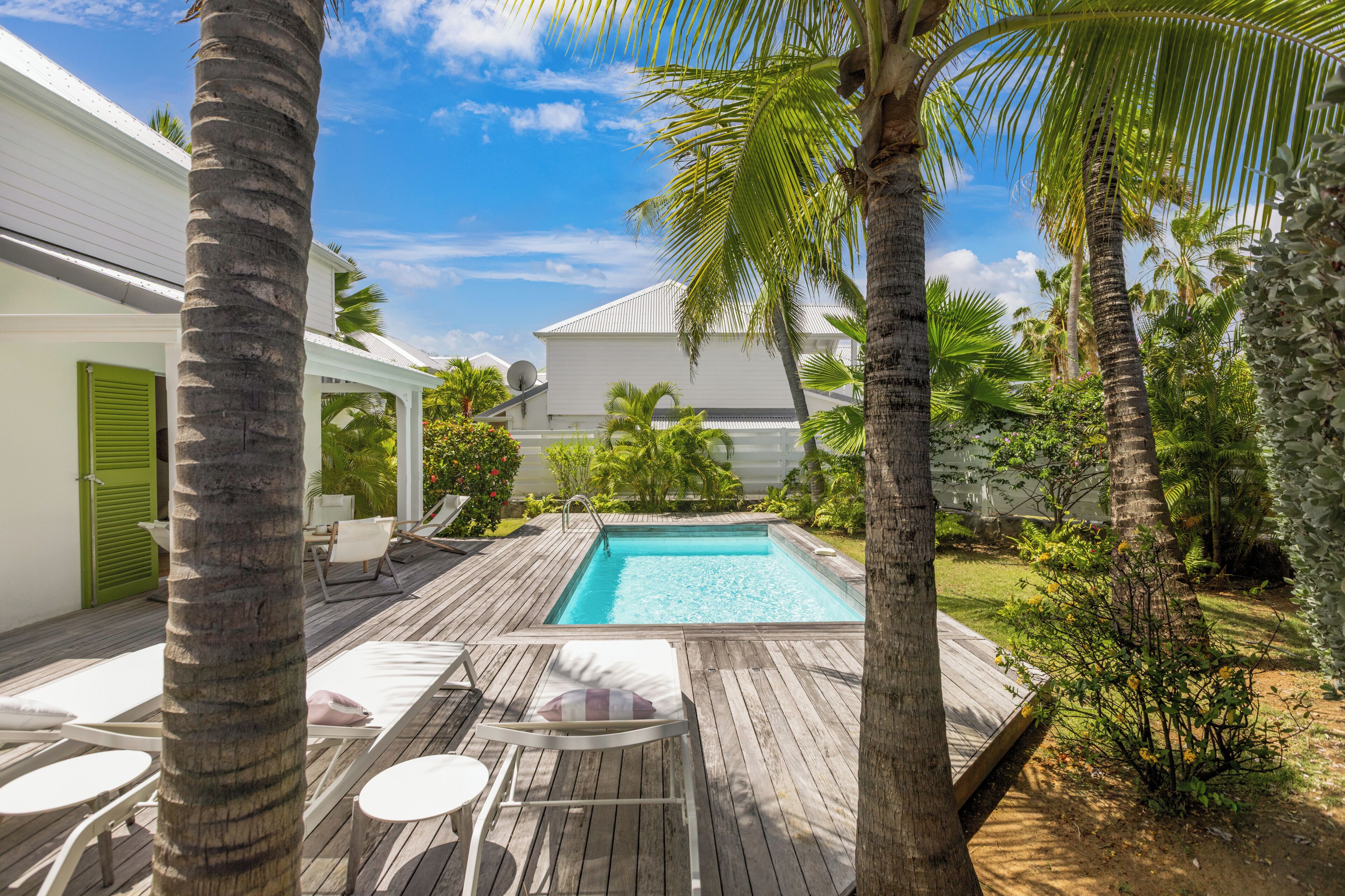 A wooden deck with lounge chairs, a swimming pool, and a white building with green shutters.