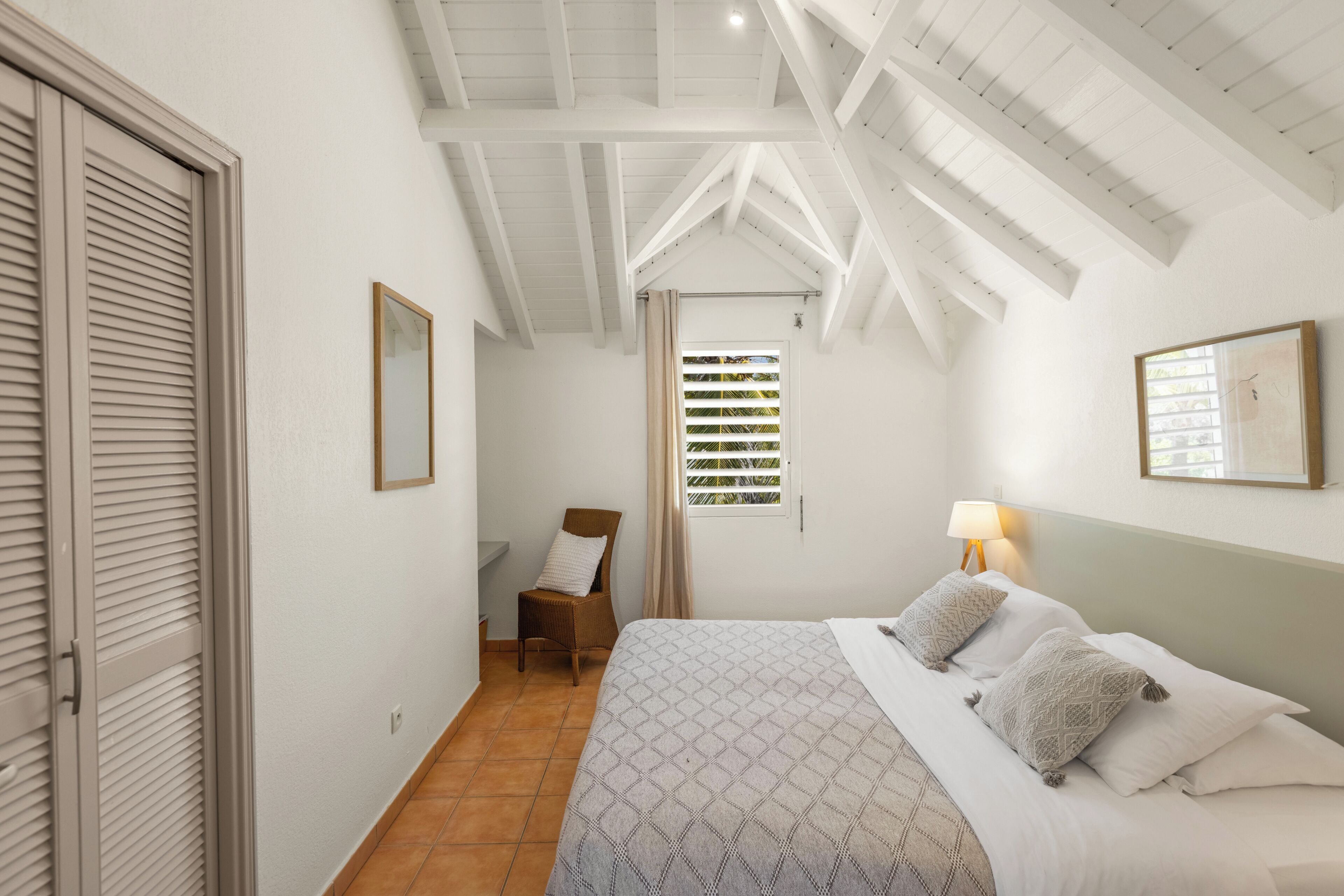 A bedroom with a bed, a chair, a window, and a ceiling with wooden beams.