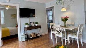 A room with a wooden floor, a dining table with chairs, a TV mounted on the wall, and a ceiling fan.