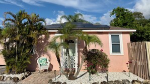 A single-story house with a sloped roof, a small porch with a chair, and a garden with palm trees and flowering plants.