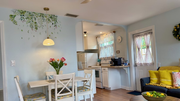 A kitchen with white cabinets, a dining table with chairs, a ceiling fan, and a window with curtains.