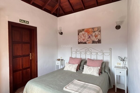 A bedroom with a bed, a wooden door labeled 'SALIDA,' a wall-mounted painting, and a white nightstand.