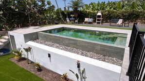 A rectangular swimming pool with a stone border, surrounded by a white wall and a grassy area with potted plants.