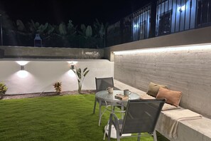 Un patio con una mesa redonda de vidrio, dos sillas, un banco acolchado y un pequeño jardín con plantas.