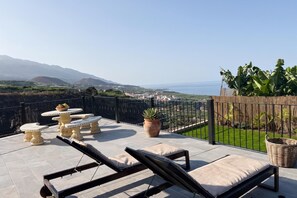 A terrace with sun loungers, a table with fruit, and a view of the sea and mountains.