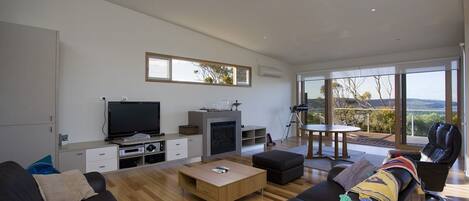 A modern living room with a flat-screen TV, fireplace, and large windows offering a view of the outdoors.