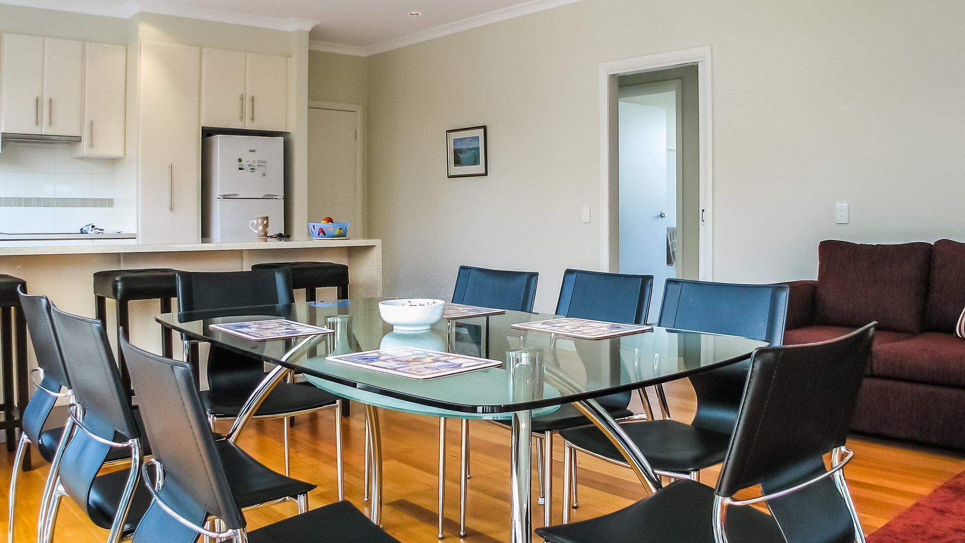 Uma sala de estar moderna com uma mesa de jantar de vidro cercada por cadeiras pretas, um sofá vermelho e uma área de cozinha com armários brancos e uma geladeira.