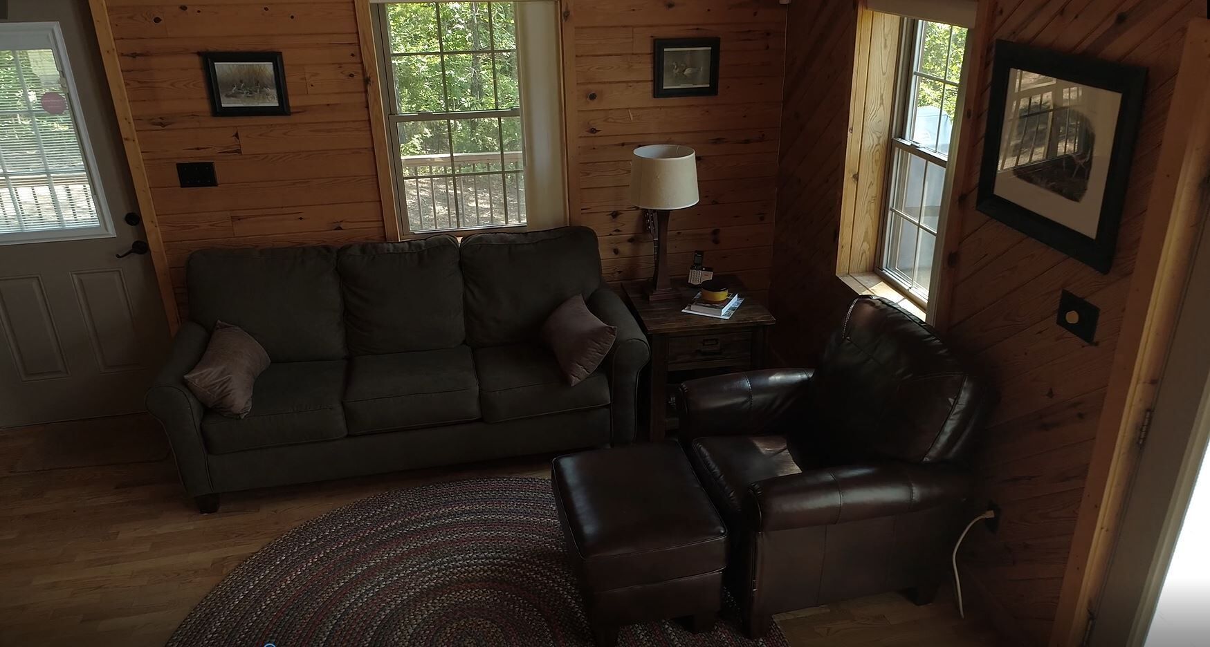 A cozy room with a wooden interior, featuring a sofa, a leather chair, a small table with a lamp, and a window with a view of trees.