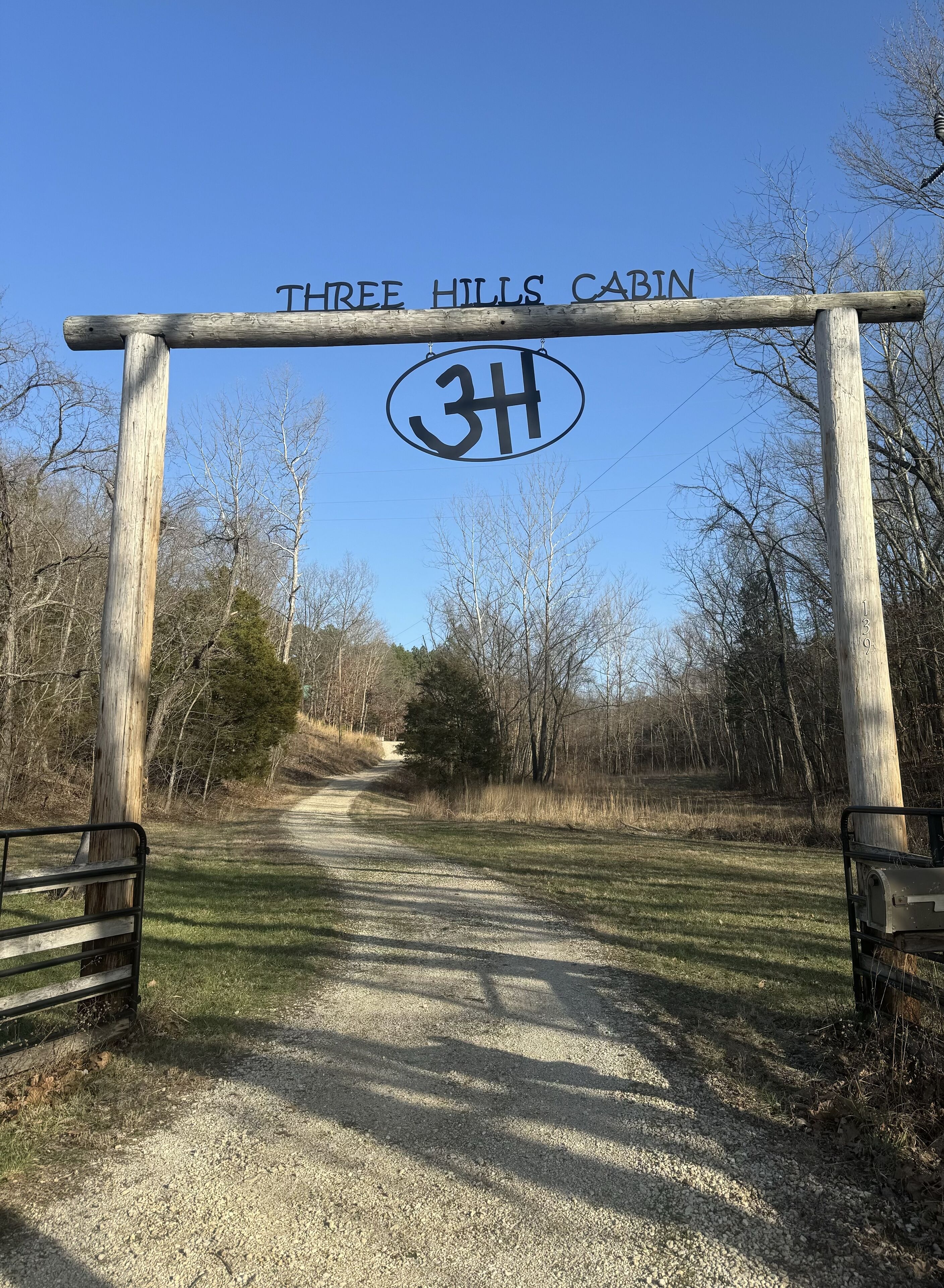 A wooden archway with "THREE HILLS CABIN" and "3H" signage, leading to a gravel path surrounded by trees.