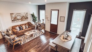 Een woonkamer met een bruine bank, een houten salontafel en een vloerkleed. Er is een deur en ramen met zonwering.