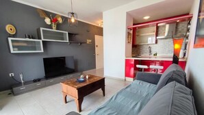 A modern living room with a flat-screen TV, a wooden coffee table, and a grey sofa. There is a kitchen visible in the background with red accents and white bar stools.