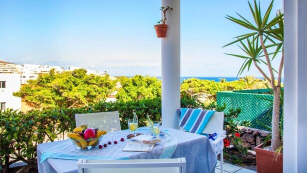 A table set with a fruit bowl, glasses, and a book, overlooking a scenic view with buildings and greenery.