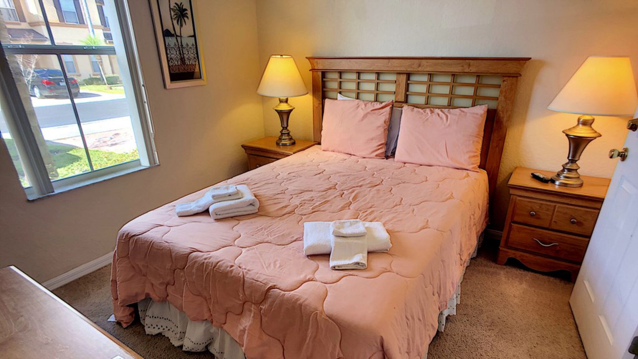 Una cama bien hecha con ropa de cama rosa, dos lámparas de noche y una ventana con vistas al exterior.