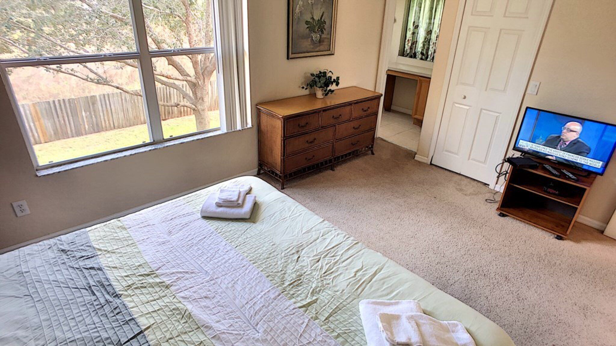 Een slaapkamer met een groot bed, een dressoir, een televisie en een raam met uitzicht op een boom en een hek.