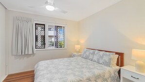 A bedroom with a bed, two bedside tables with lamps, a ceiling fan, a window with curtains, and a view of buildings outside.