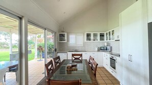 A kitchen with white cabinets, a dining table with chairs, and a view through the glass door to the outside. - Riverside - Burrum Heads- Riverfront - 3BR - River Fishing... (BURRUM HEADS)
