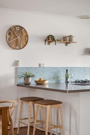 A kitchen with a wooden clock, a plant, a shelf with kitchen items, and two stools.