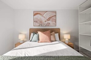 A hotel room with a bed, two bedside tables with lamps, a large abstract painting above the bed, and a shelving unit to the right.