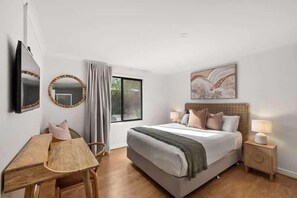 Un dormitorio con cama, mesitas de noche, escritorio, silla, ventana y televisor montado en la pared.