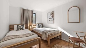 A bedroom with two beds, a chair, a window with curtains, and a wall-mounted mirror. - Cormorant Cottage—Garden Beach Abode w/ Game Room (Dunsborough)