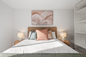Un dormitorio ordenado con cama, dos mesitas de noche con lámparas, una obra de arte en la pared y una estantería.