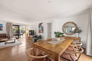 Een ruime woonkamer met een houten eettafel, rieten stoelen en een grote spiegel.
