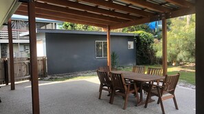 An outdoor patio with a wooden table and chairs, a pergola overhead, and a view of a house with a dark exterior wall and a window.