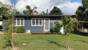 A single-story house with a dark exterior, white-framed windows, and a front door with a glass panel. There is a small porch area with a wooden deck, and the property is surrounded by a well-maintained lawn and various plants. - 9 Indigo Avenue - Rainbow Beach. Close to beach and main street. Walk to everything (RAINBOW BEACH)