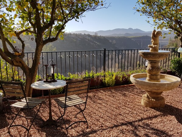 Une terrasse avec une table et des chaises, une fontaine en pierre et une vue sur une vallée bordée de montagnes.