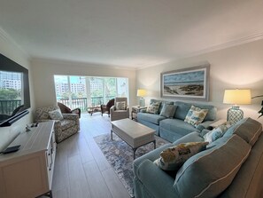 A modern living room with a sofa, armchairs, a coffee table, and a TV. There is a balcony with a view of buildings and trees.