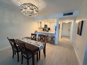 A dining area with a table and chairs, a kitchen with white cabinets, and a hallway leading to a door.