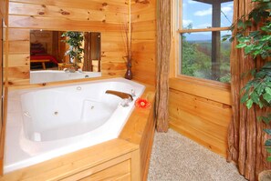 Un baño de cabaña de madera con una bañera grande, un espejo y un ventanal con vistas a un bosque.