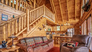 A log cabin interior with a wooden staircase, a leather sofa, a dining table, and a kitchen area.