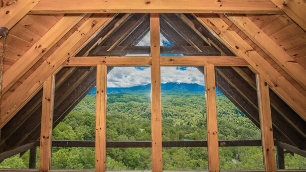 A wooden attic room with a large window offering a view of a forested valley.