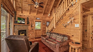 A log cabin interior with a fireplace, a leather chair, a sofa with patterned cushions, and a wooden staircase.