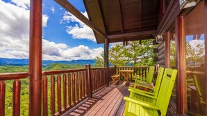 A wooden deck with a view of green mountains, a wooden railing, and two green chairs.